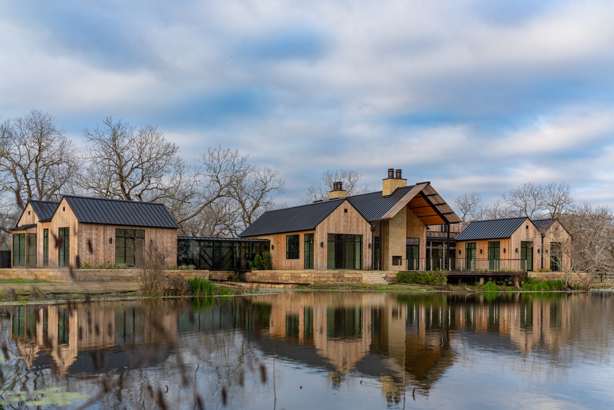 View from far away across the pond looking back at the Fulshear Modern Ranch Estate featuring Kebony Modified Wood Character Decking in Fulshear, Texas on a cloudy evening