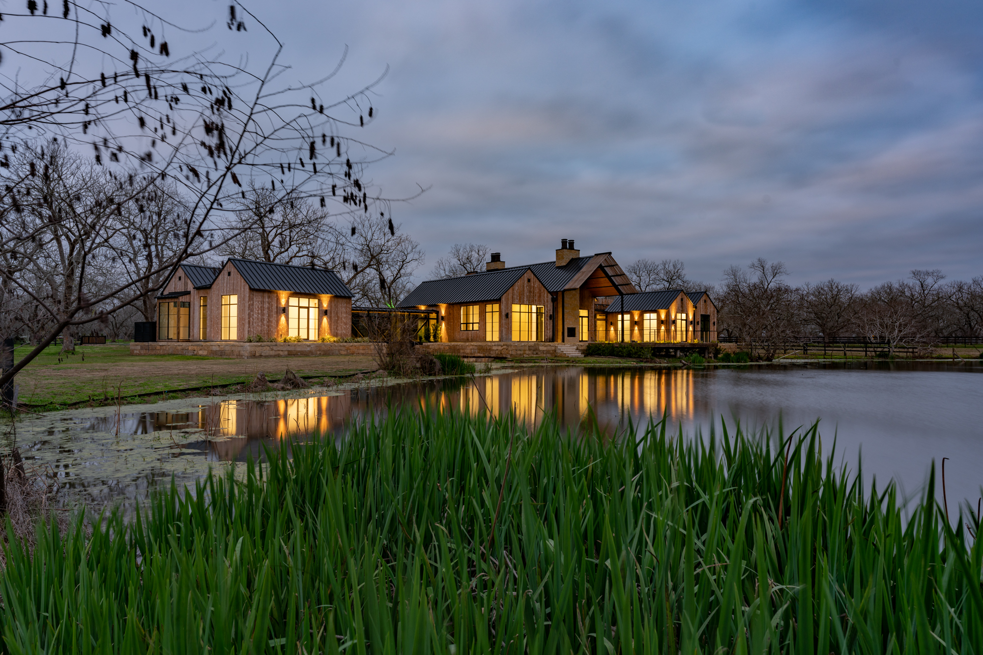 View at night from far away across the pond looking back at the glowing interior of the Fulshear Modern Ranch Estate featuring Kebony Modified Wood Character Decking in Fulshear, Texas