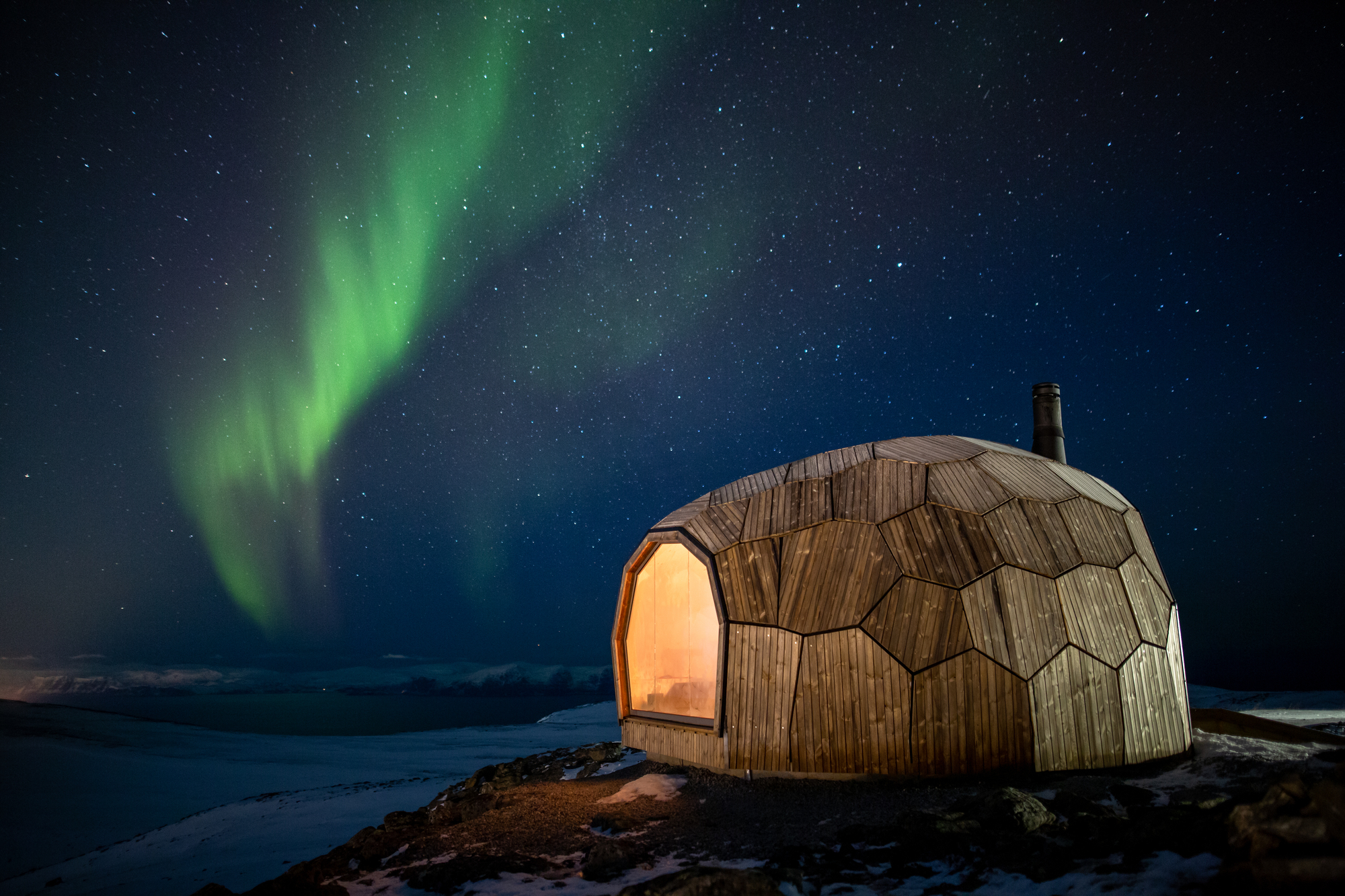 The aurora borealis illuminates the surrounding snow in the nighttime sky above a lit up Hammerfest Cabin nestled in Hammerfest, Norway and features Kebony Modified Wood Character Cladding