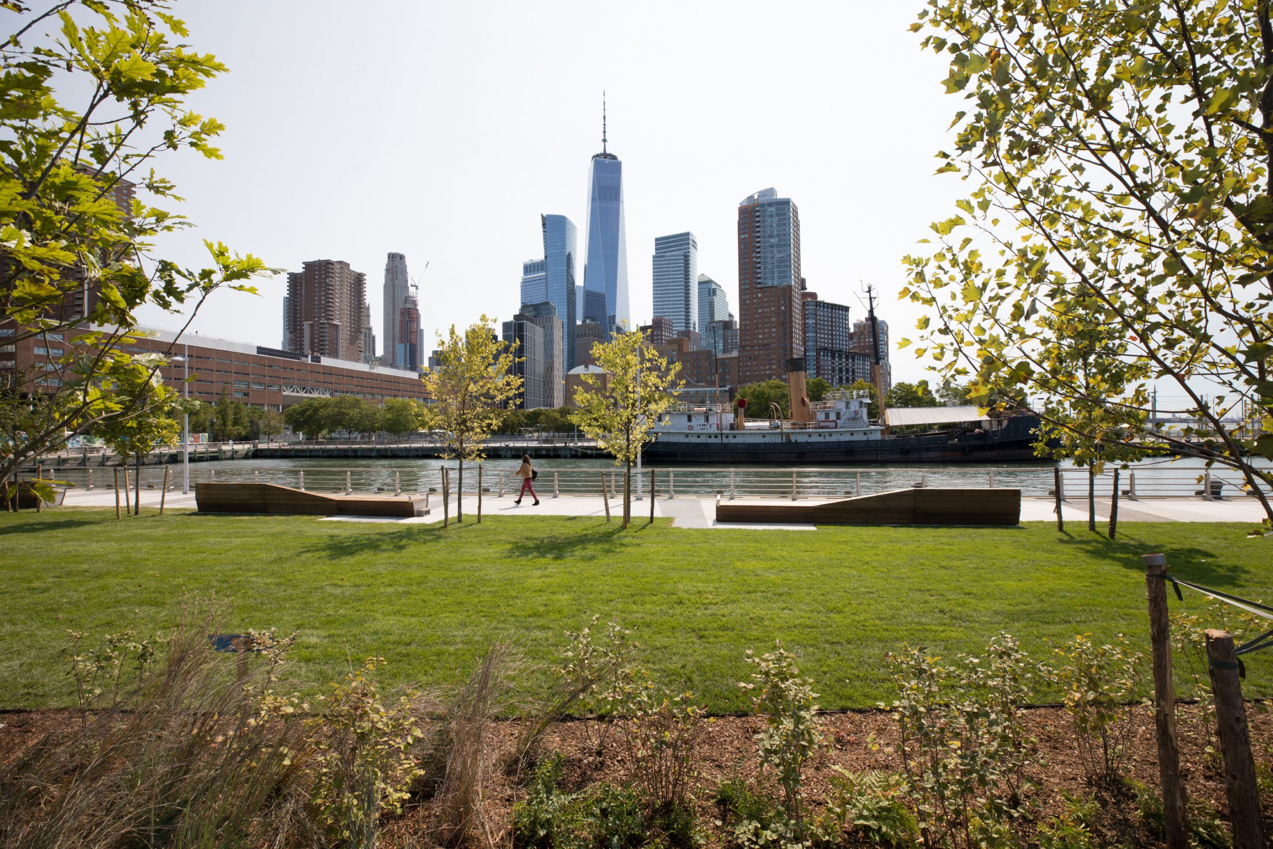 Looking at Pier 26 featuring kebony modified wood Clear Boardwalk Decking from the side with nature and trees in the foreground during the day at Hudson River Park in New York City New York. The downtown skyline is in the background.
