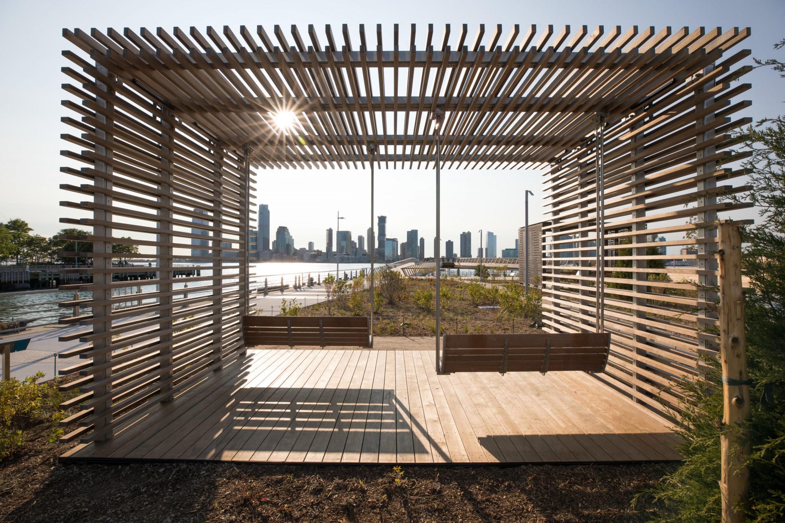 Two bench swings with a slatted roof over look the Hudson River at Pier 26 during the day at Hudson River Park in New York City New York features kebony modified wood Clear Boardwalk Decking.
