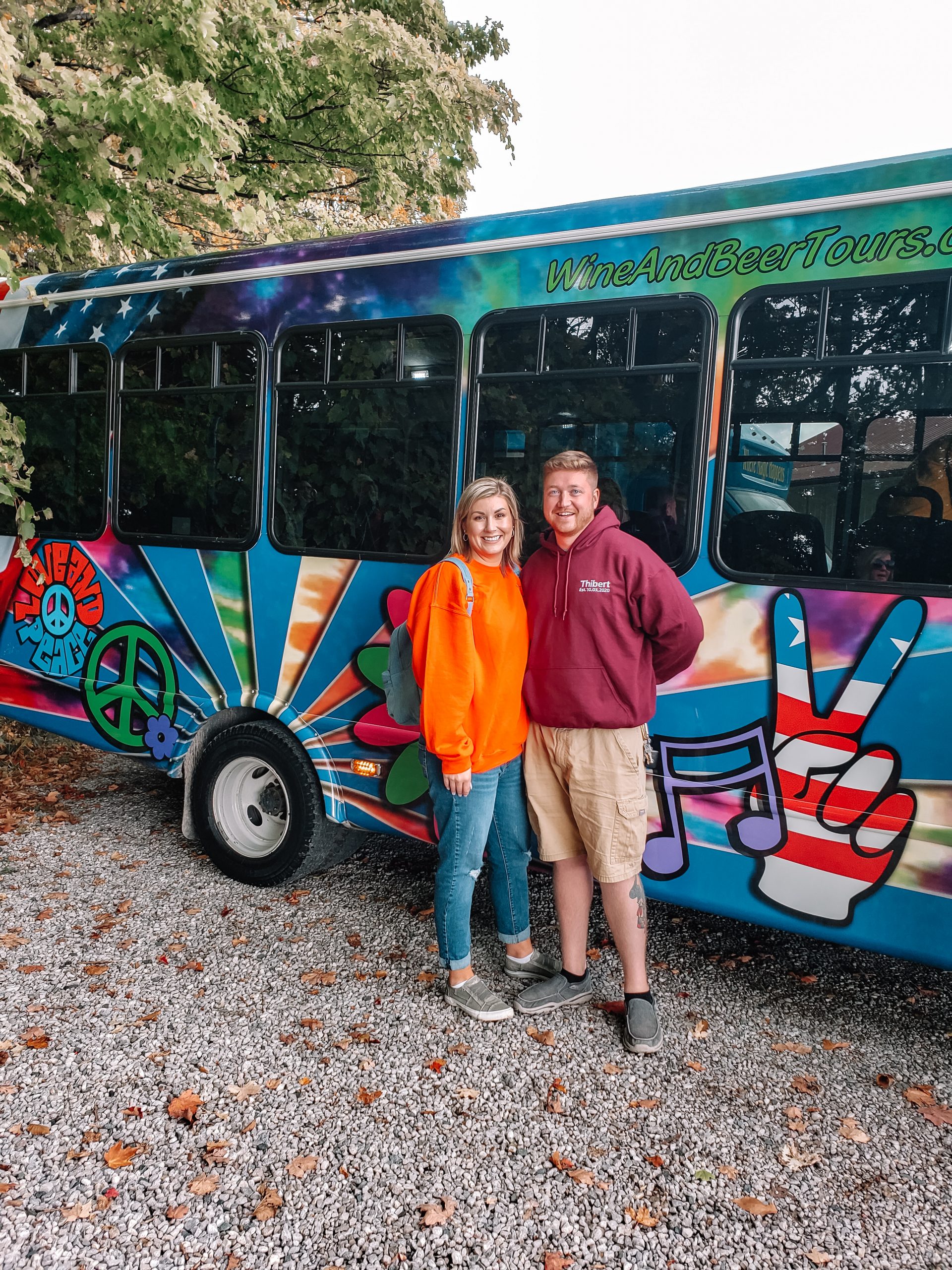 Kelsey Thibert and her husband smile in front of a colorful tour bus on a sunny day.