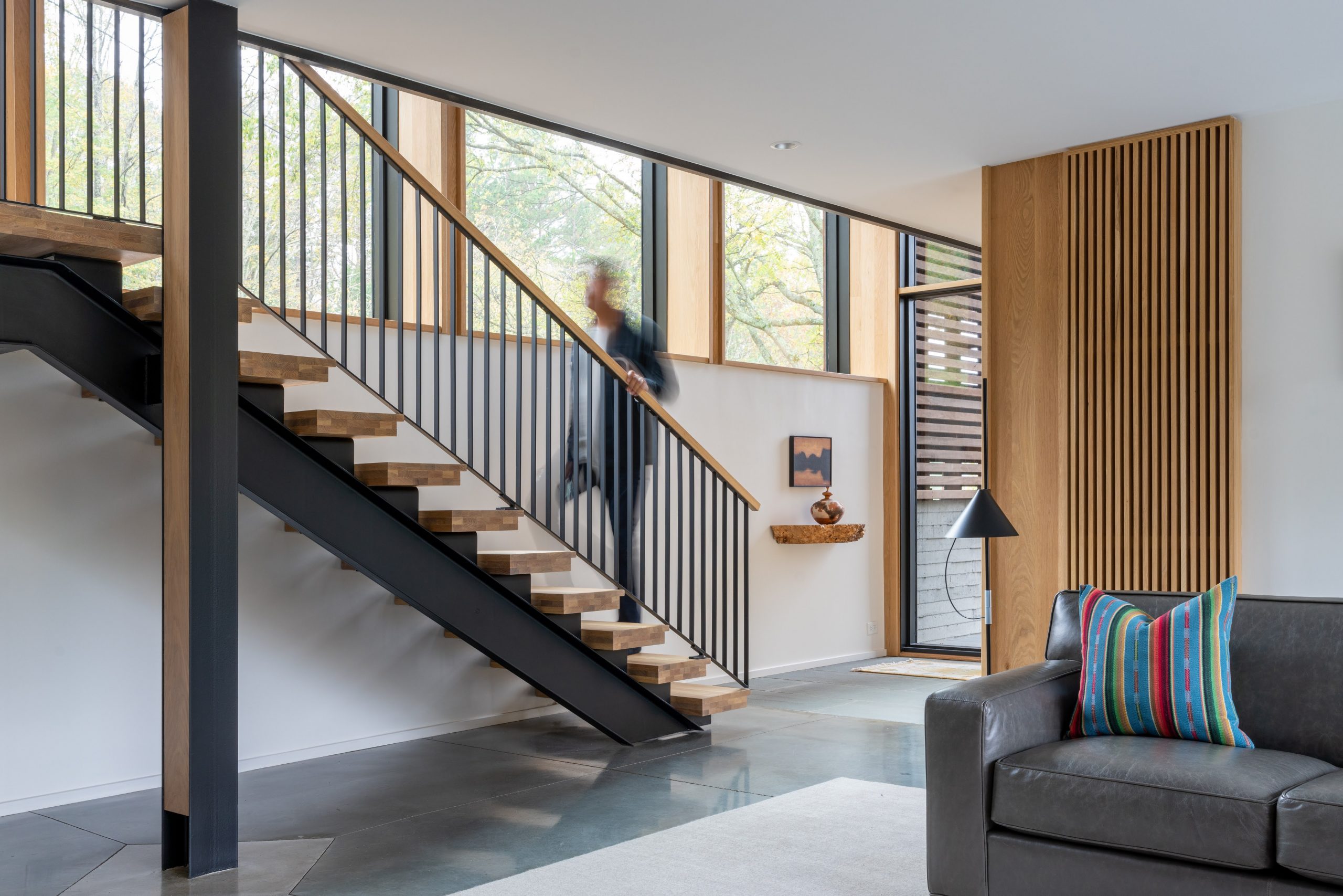 A person walks up the stairs on the side of the living room of the Wood Screen House in Nashville, Tennessee during the day featuring Kebony Modified Wood Clear Cladding on the siding of the back wall and the edge of a gray couch in the foreground on the right