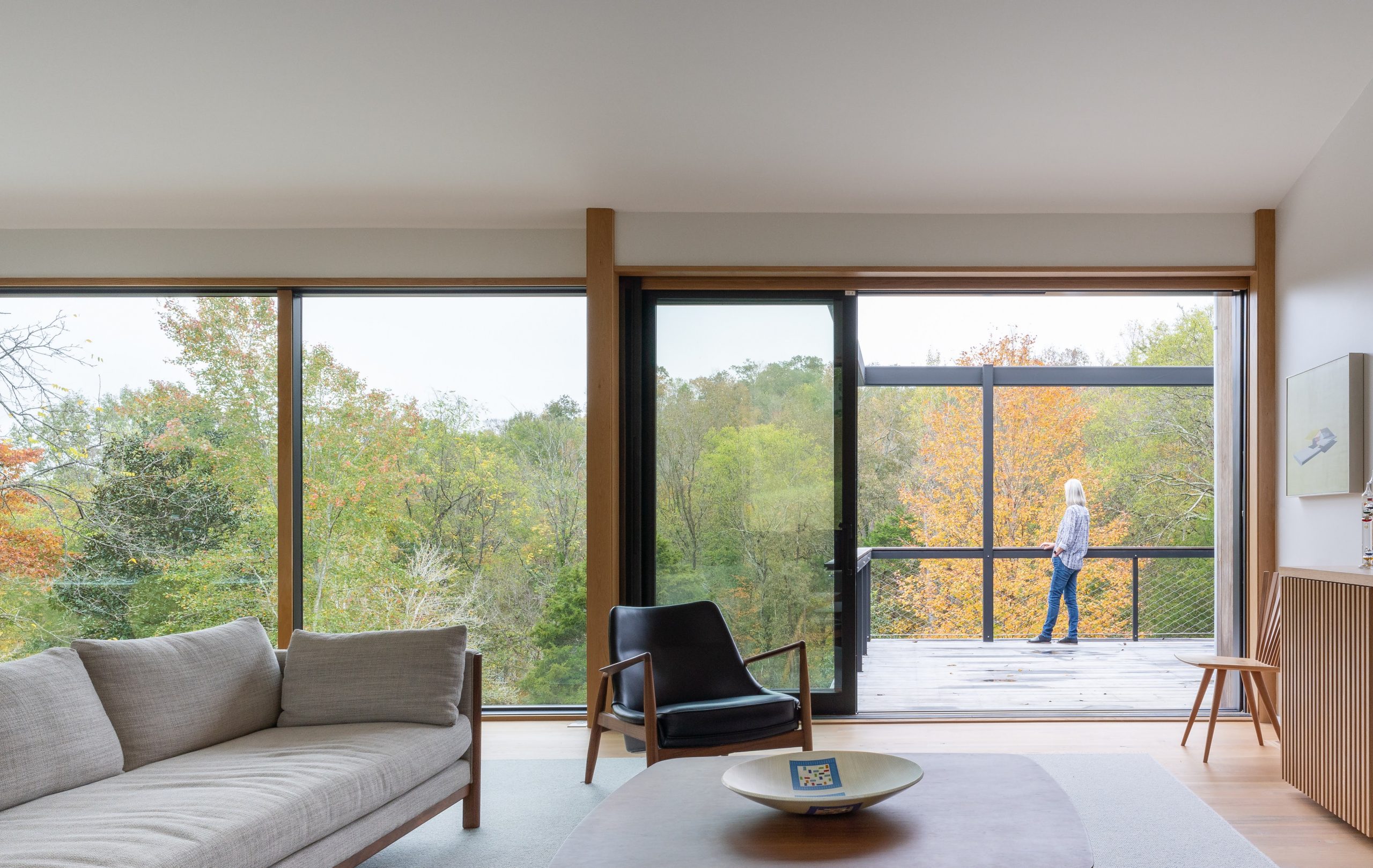 Interior view of the second story living room with lavish monochrome furnishings and a person on the outside balcony looking at the woods of the Wood Screen House in Nashville, Tennessee during the day featuring Kebony Modified Wood Clear Cladding on the siding