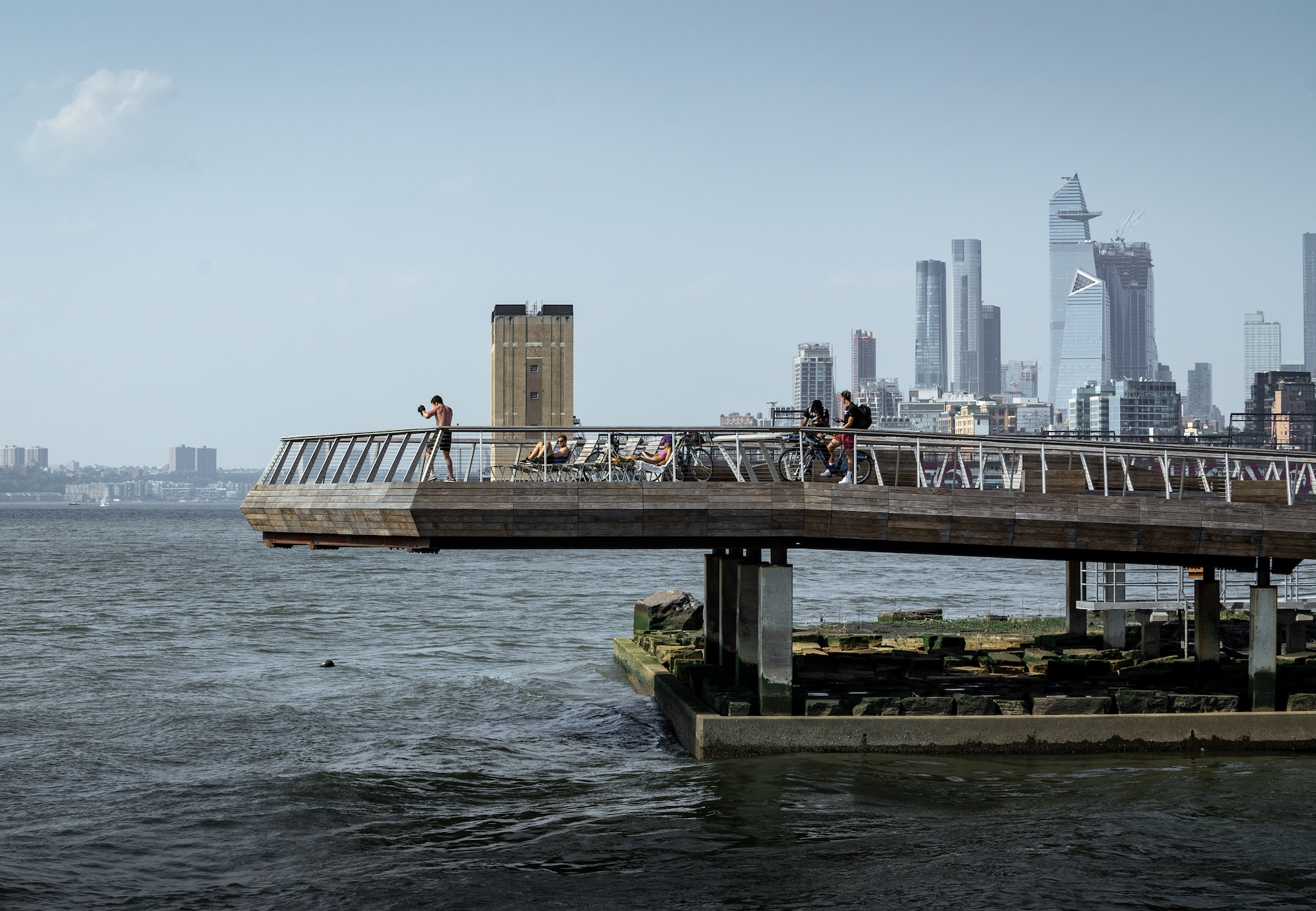 Side view of people enjoying The Tide Deck at Pier 26 featuring kebony modified wood Clear Boardwalk Decking during the day with the city skyline in the background at Hudson River Park in New York City New York Downtown New York City