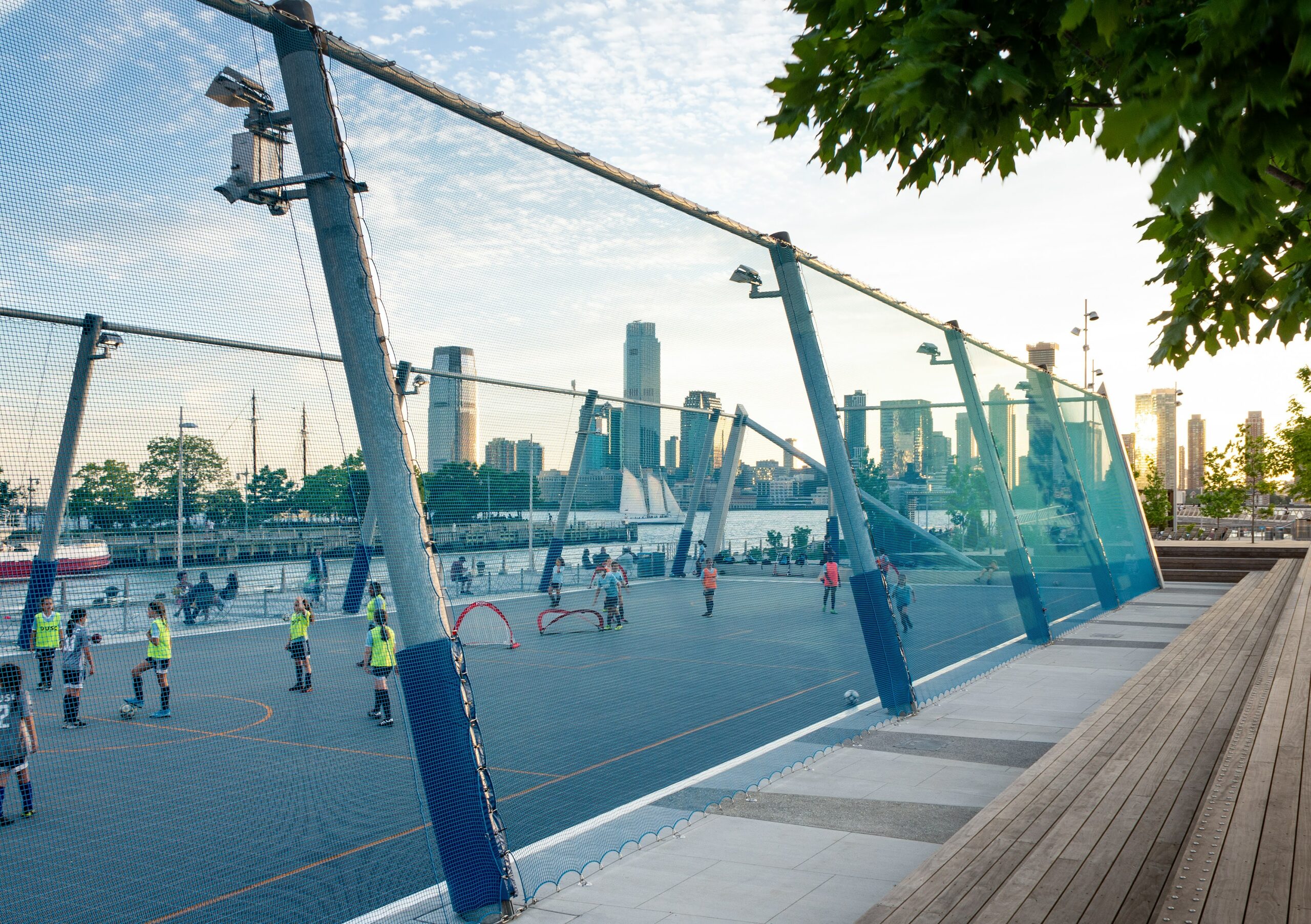 Side view of a fenced off area for children's sports at The Tide Deck at Pier 26 featuring kebony modified wood Clear Boardwalk Decking during a sunset with the city skyline in the background at Hudson River Park in New York City New York Downtown New York City
