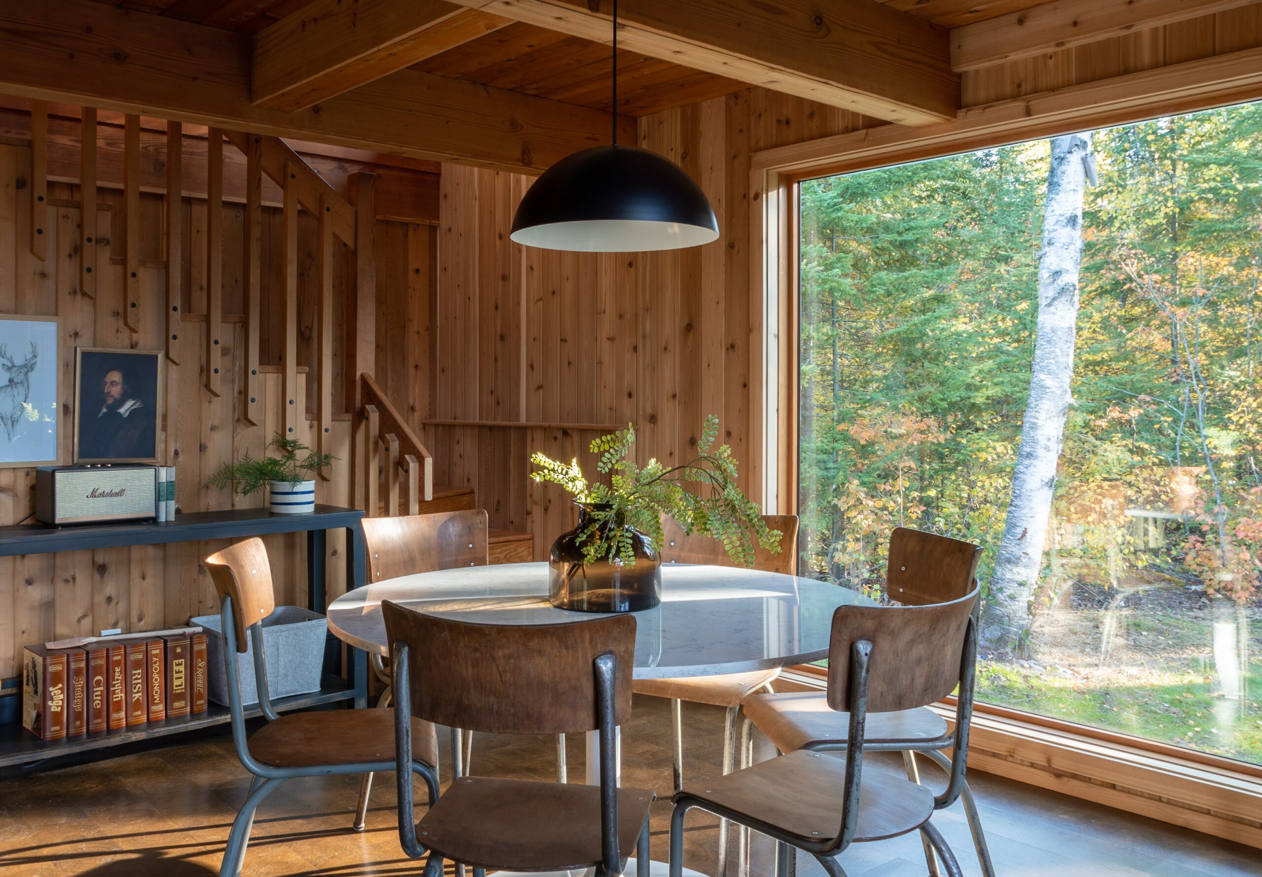 Interior view of a circular dining room table of The Minne Stuga Cabin in Grand Marais Minnesota prominently featuring Kebony modified wood siding throughout the houses' design with a giant floor to ceiling window showing the woods outside.